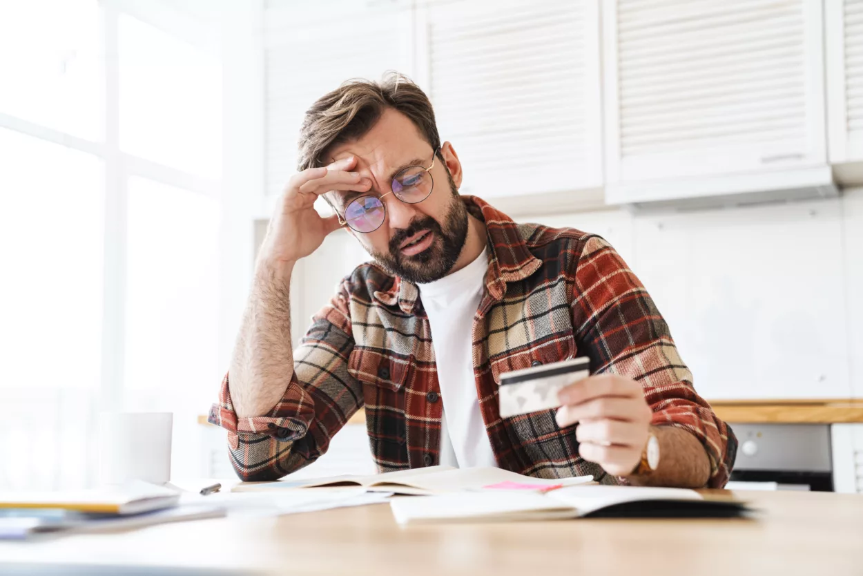 A man in glasses, wearing a plaid shirt, looks concerned while holding a credit card in a kitchen. Documents and a coffee mug are on the table.
