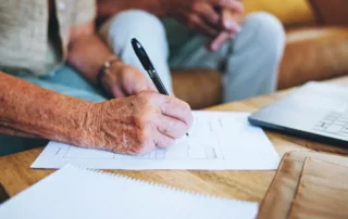 Elderly person writing on a document with a pen at a wooden table, with an open laptop nearby, conveying focus and attentiveness in legal planning.