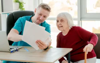 Healthcare professional in blue scrubs assisting an elderly woman with a cane, reviewing documents at a desk in a warm, supportive setting.