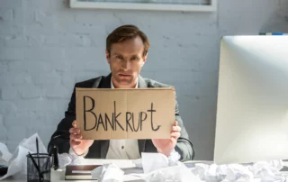 Man holding “Bankrupt” sign at cluttered desk, representing Chapter 7 bankruptcy, financial stress, and concerns about what assets can be kept during filing